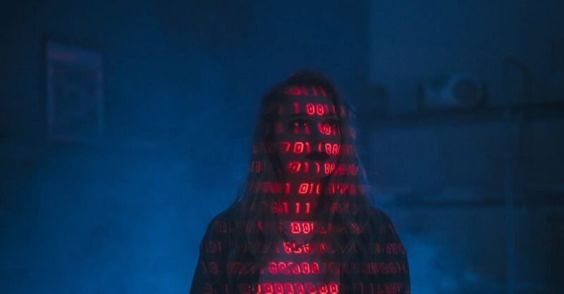 A mysterious silhouette with red binary code projected over the face, set against a dark, moody background.