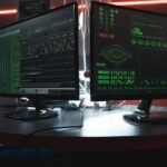 Close-up of dual computer monitors with green coding interfaces in a dark room, highlighting cyber security themes.
